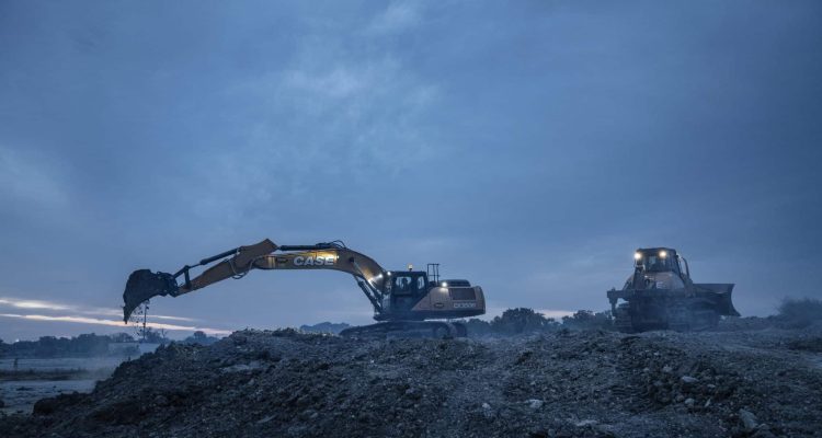 A CASE backhoe and bulldozer operating on a construction site after sunset with a cloudy sky background and low light.