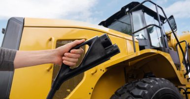 A closeup of a yellow wheel loader and a person holding a black EV charger in front of it.