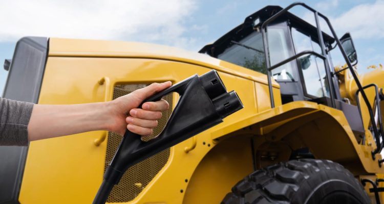 A closeup of a yellow wheel loader and a person holding a black EV charger in front of it.