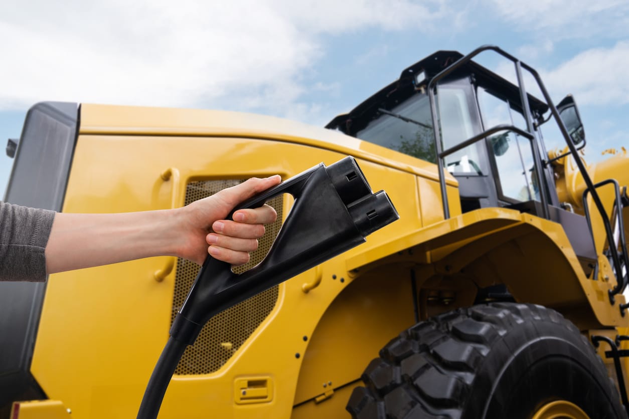 A closeup of a yellow wheel loader and a person holding a black EV charger in front of it.
