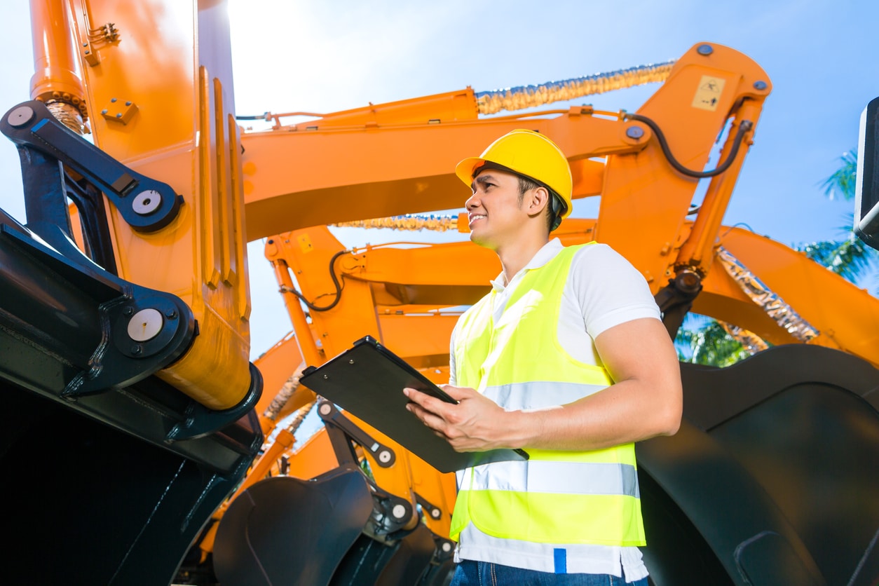 A construction worker in a neon vest and yellow hard hat holding a clipboard during a construction site safety inspection.
